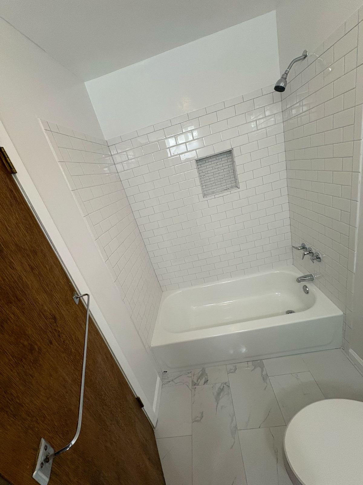 Completed bathroom with white subway wall tile, bathtub, shower fixtures, recessed tile niche and light patterned floor tile. Client project photo; exact job location is not documented.