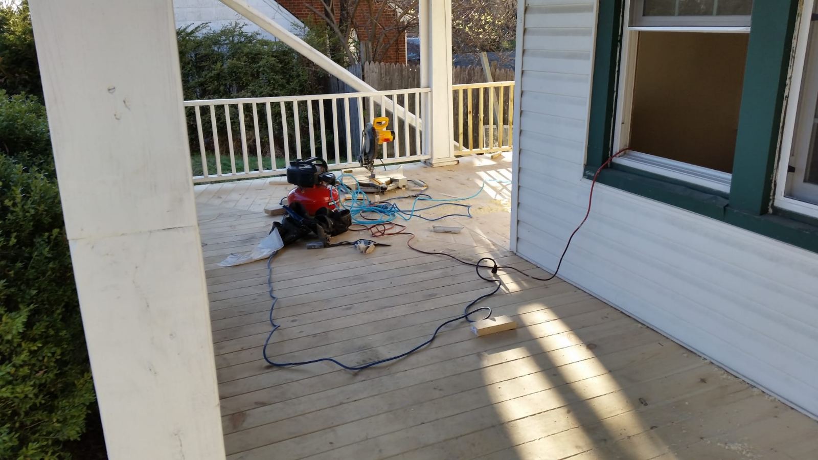 Porch and deck carpentry work in progress, showing wood floor boards, white railings and posts, siding and repair tools. Client project photo; exact job location is not documented.